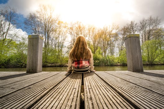 A young girl sits on a wooden dock, enjoying a sunny day by the water surrounded by trees.